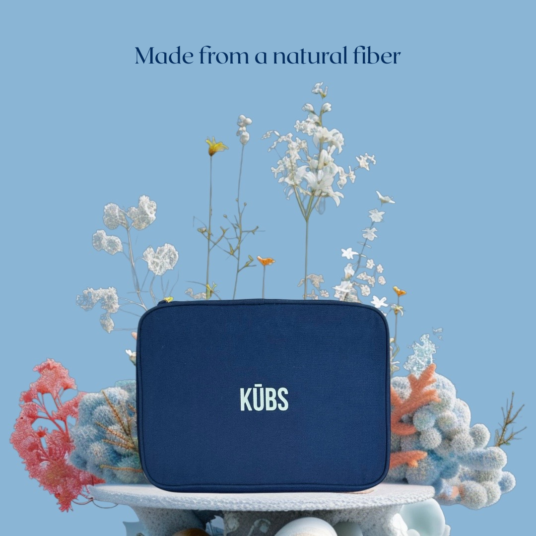 Eco-styled KŪBS cotton packing cube displayed on a textured coral-like platform, surrounded by delicate dried and natural flowers. Emphasizes sustainable materials. Light blue background with dark navy cube. Available in Canada and USA.